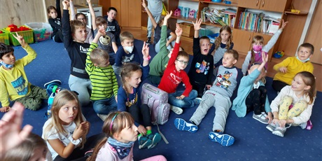 Powiększ grafikę: Uczniowie siedzą w grupie i słuchają nauczycieli świetlicy, część z nich podnosi rękę.